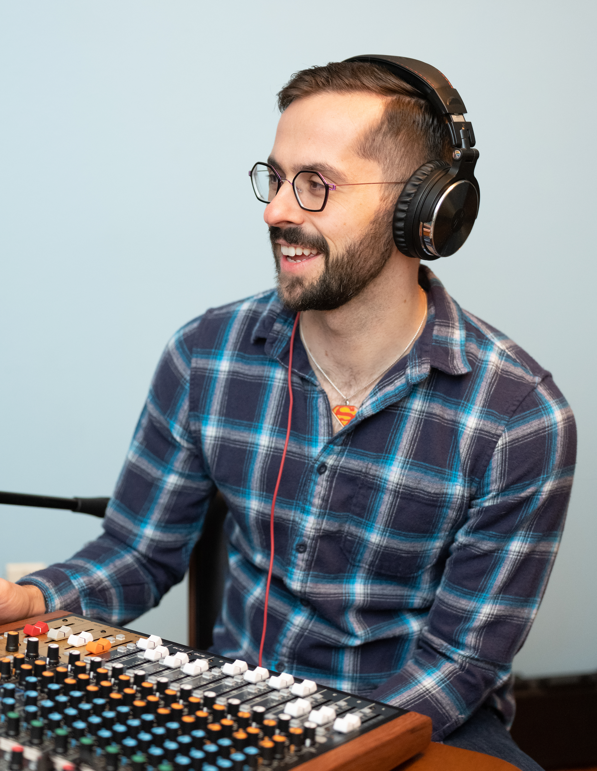 Derek is sitting in front of a soundboard wearing headphones. He's smiling and looking off camera. He wears a blue plaid button-down shirt.