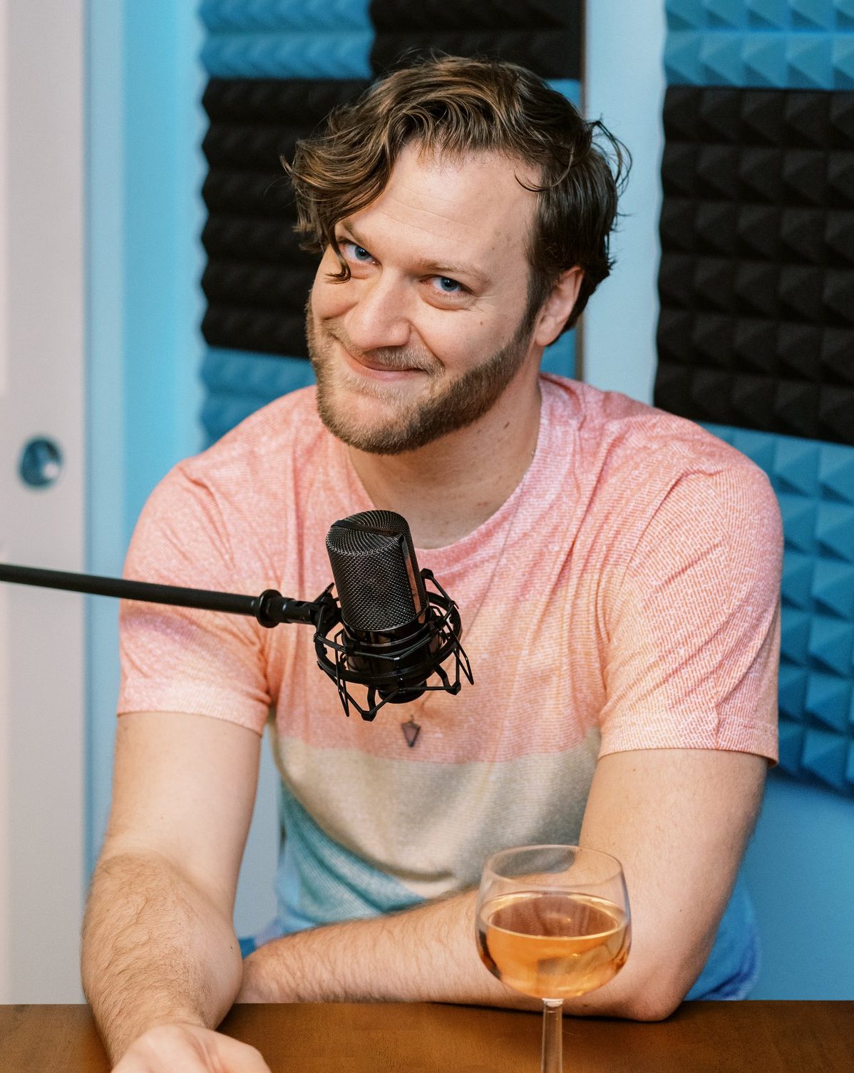 Kyle is sitting at a table smirking at the camera. He is wearing a rainbow shirt with a mic in front of him and a glass of rose on the table.